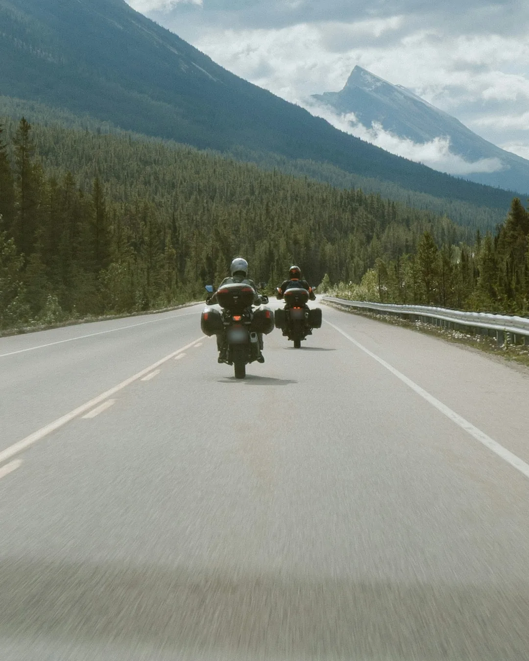 Two riders on touring motorbikes riding together on an open road through pine forest, toward the mountains